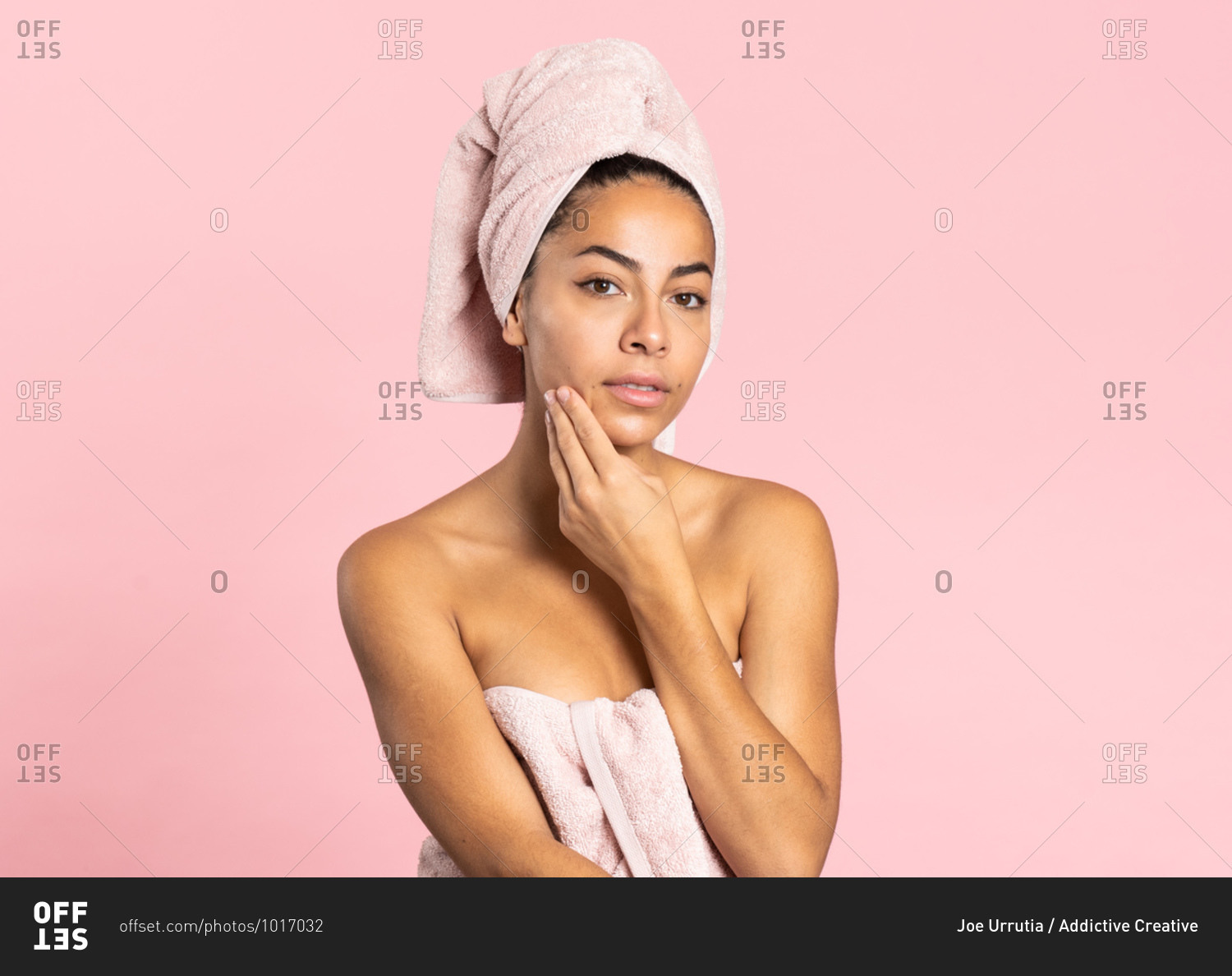 Charming young ethnic bare shouldered female with bath towel on head and perfect olive skin looking at camera touching skin standing against pink background while representing beauty and skincare concept