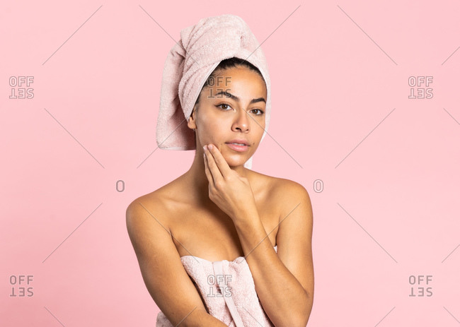 Charming young ethnic bare shouldered female with bath towel on head and perfect olive skin looking at camera touching skin standing against pink background while representing beauty and skincare concept