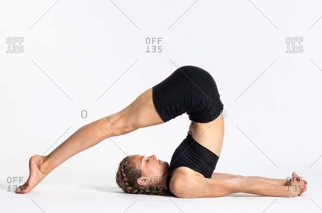 Full body side view of young slim female in sportswear doing Plow yoga asana against white background