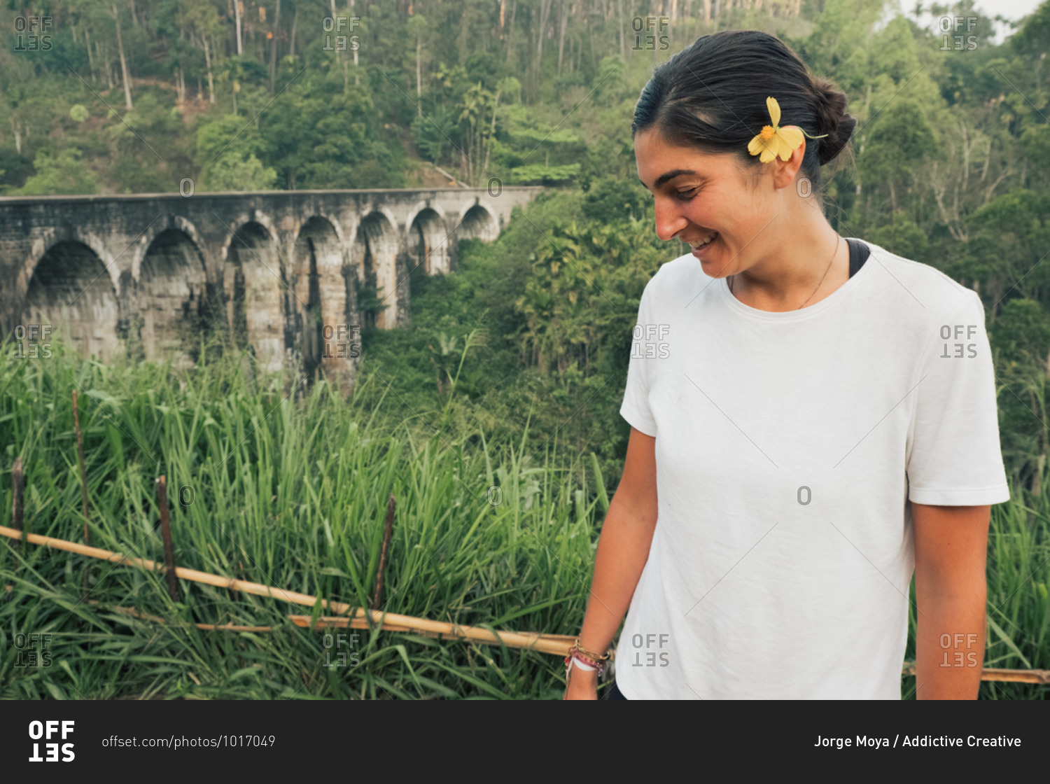 Smiling young ethnic female traveler with yellow flower in hair resting in green tropical forest near old arched bridge in Sri Lanka