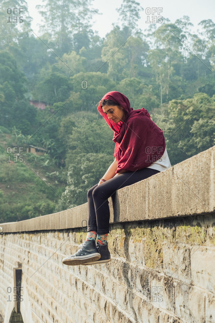 Low angle side view of thoughtful young ethnic female traveler in active wear and headscarf sitting on stone border of aged bridge against green tropical foliage during vacation in Sri Lanka