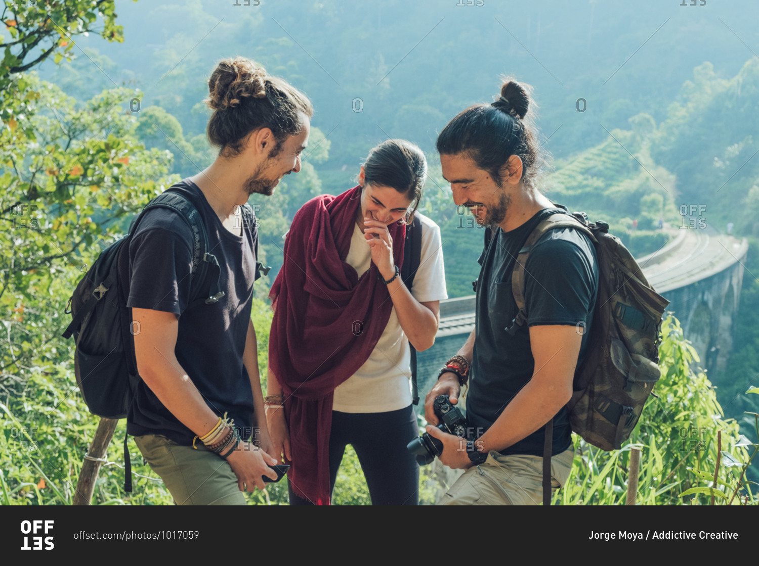 Group of cheerful young multiethnic friends with backpacks and photo camera having fun and laughing while standing on green hill near old viaduct bridge during summer holidays in Sri Lanka