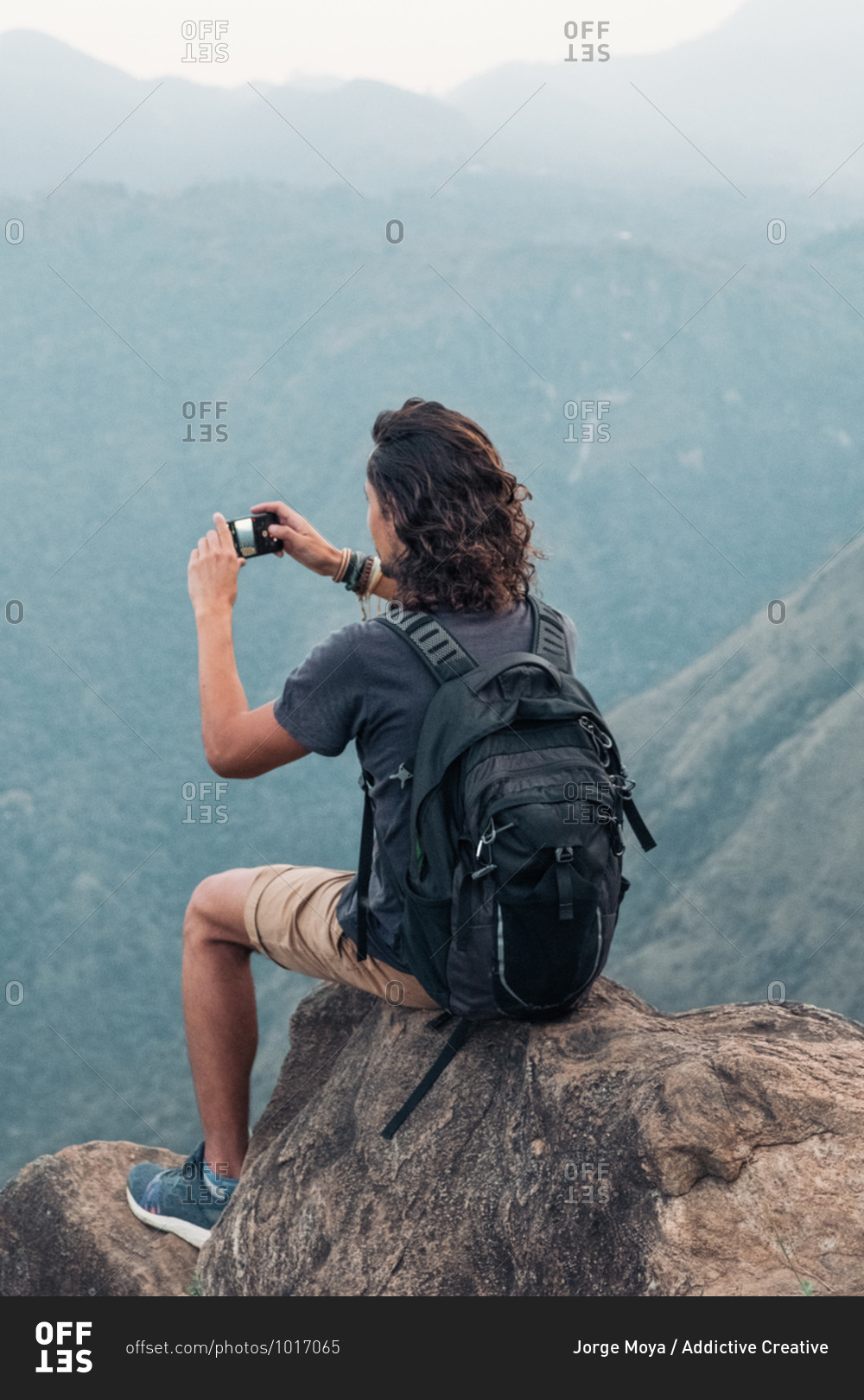 Side view of traveling male sitting on rock and taking picture on smartphone of amazing mountainous landscape in Sri Lanka