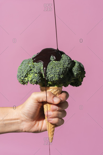 Studio shot of man's hand holding ice cream cone with broccoli on top and pouring chocolate sauce, against pink background
