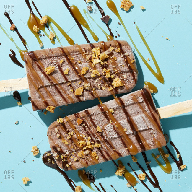 Two homemade fudge iced lollies with sprinkled and drizzled nuts, caramel and chocolate sauce on blue background, overhead view