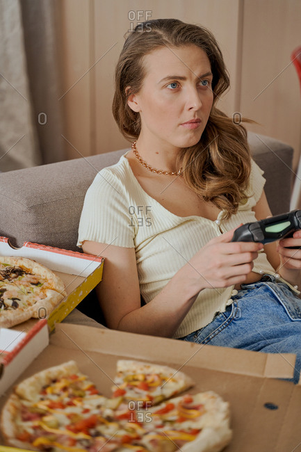 Blonde woman sitting on sofa eating pizza and playing video games