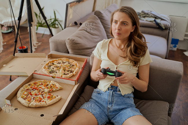 Young blonde woman sitting on sofa eating pizza and playing video games