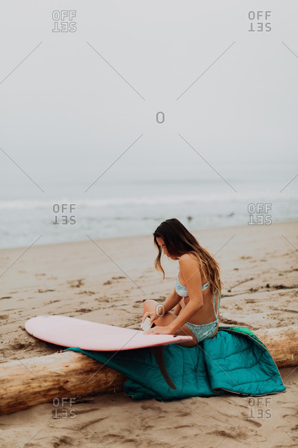 Young female surfer sitting on log waxing surfboard at beach, Ventura, California, USA