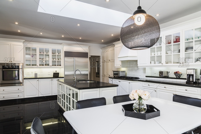 White breakfast table with black leather chairs, white wood and glass pane island with black granite countertop,  cupboards in country style kitchen, black granite tile flooring