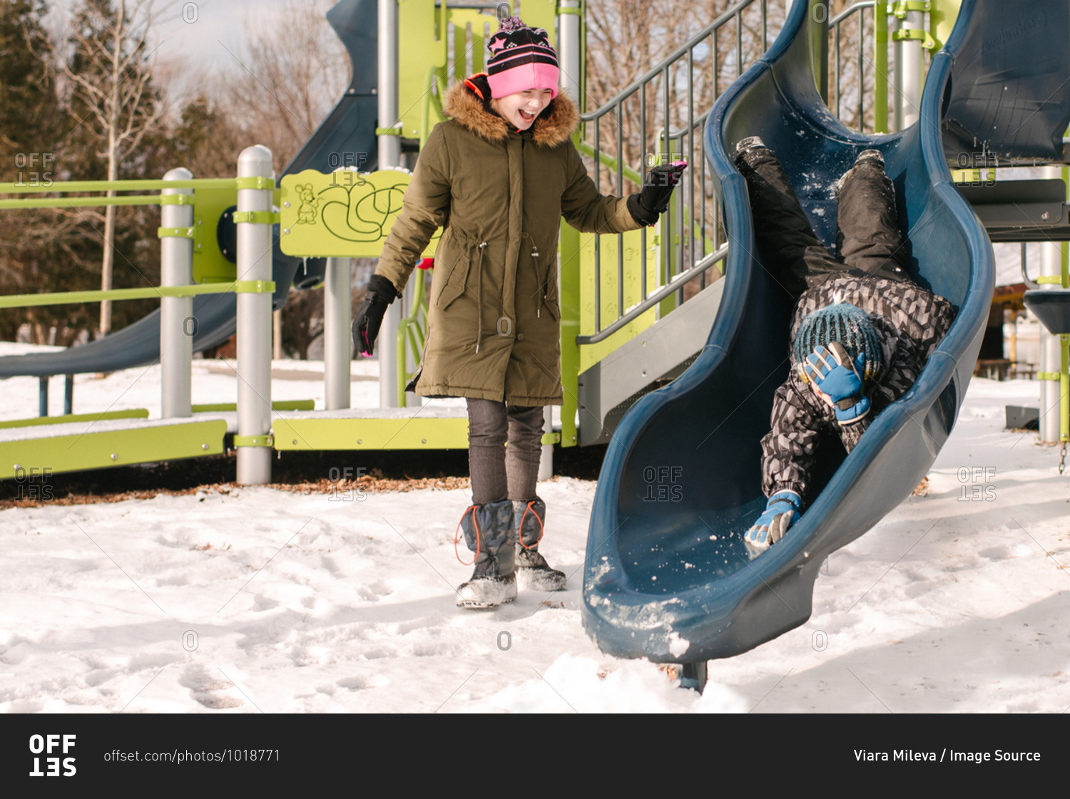 Girl watching brother headfirst on playground slide in snow