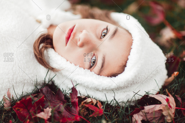 Girl in white hooded top lying on grass amongst autumn leaves, portrait