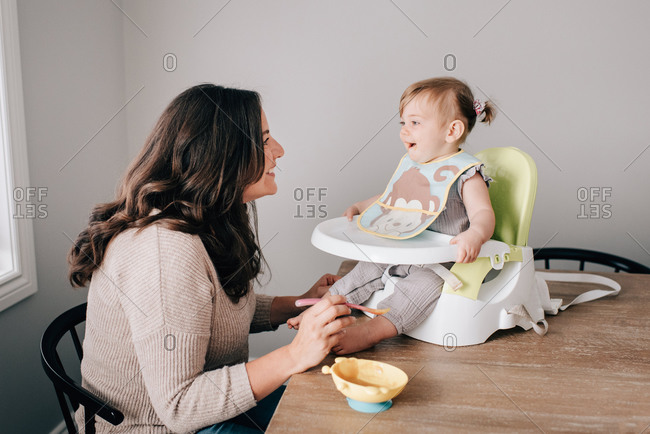 Mother feeding baby daughter in child seat on kitchen table