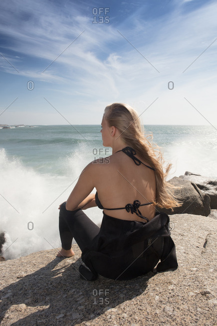 Young female surfer with long blond hair sitting on beach rock, rear view, Cape Town, Western Cape, South Africa