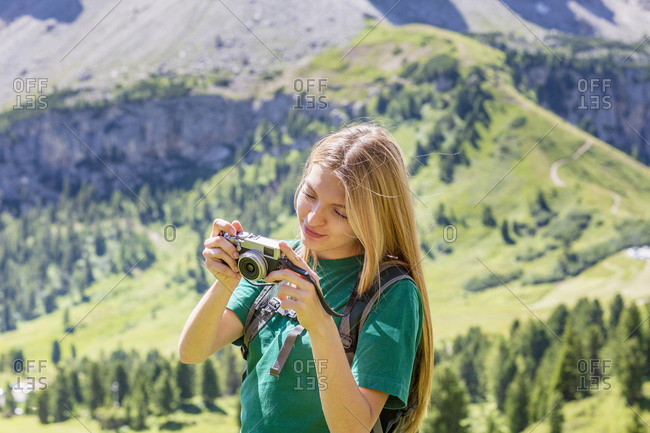 Young woman examining pictures on photo camera during summer vacation in Val Badia valley on blurred background of Dolomites mountains in Italy