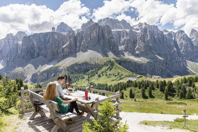 Side view of young man and woman sitting on lumber bench at table on terrace in green Val Badia valley near Dolomites mountain range on cloudy summer day in Italy