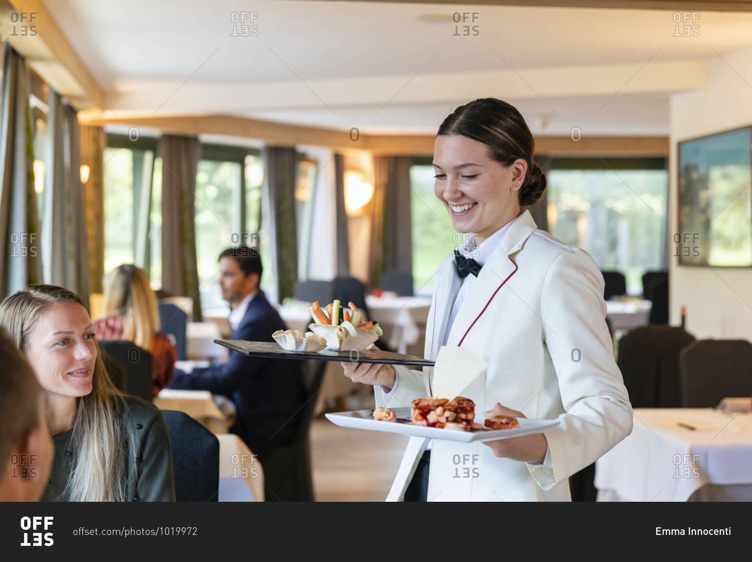 Positive woman in uniform smiling and carrying plates with exquisite food for clients while working in cozy restaurant