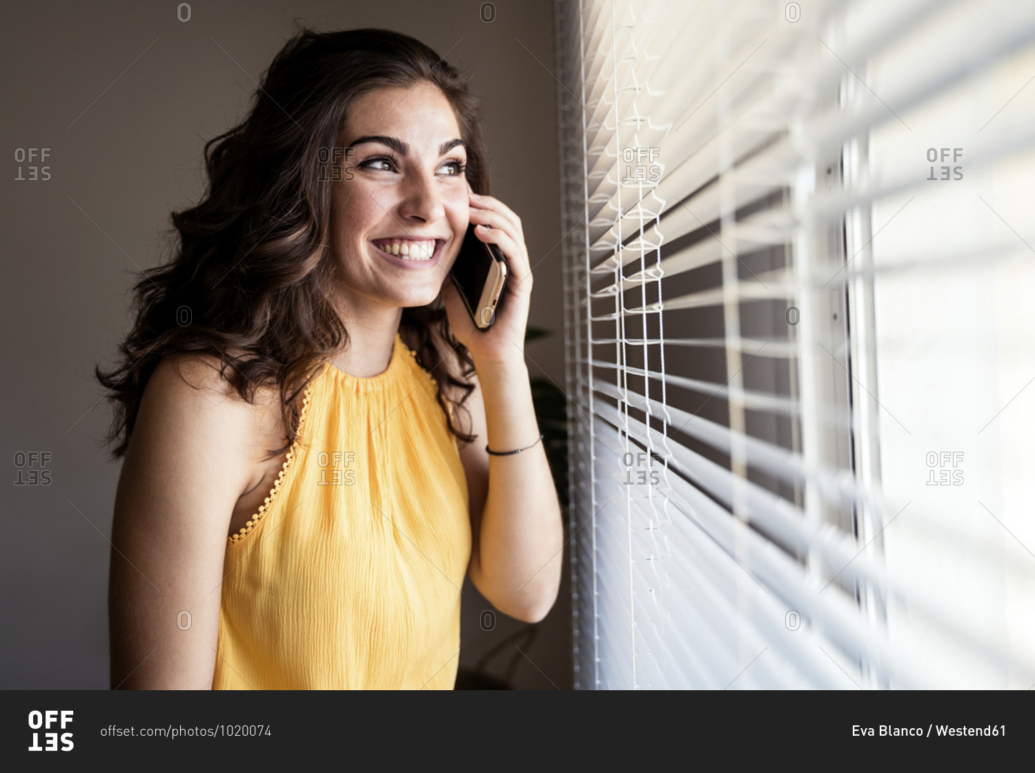 Cheerful young woman talking on smart phone while looking away through blinds at home