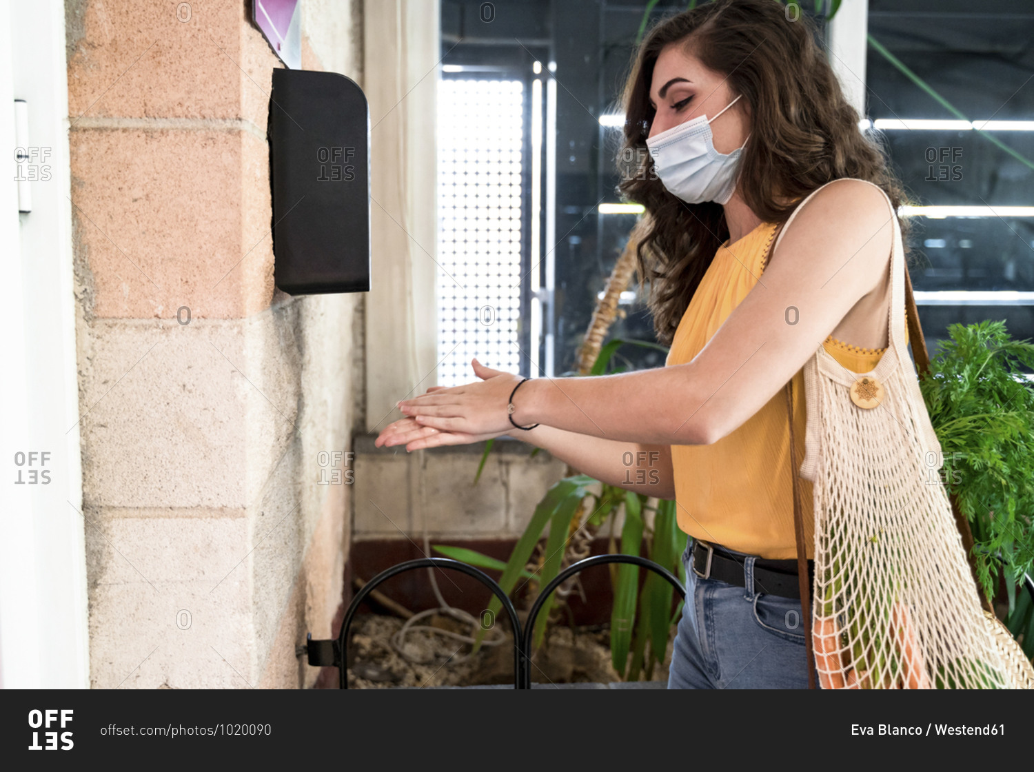 Young woman wearing face mask rubbing hands with alcohol disinfection gel by dispenser on wall