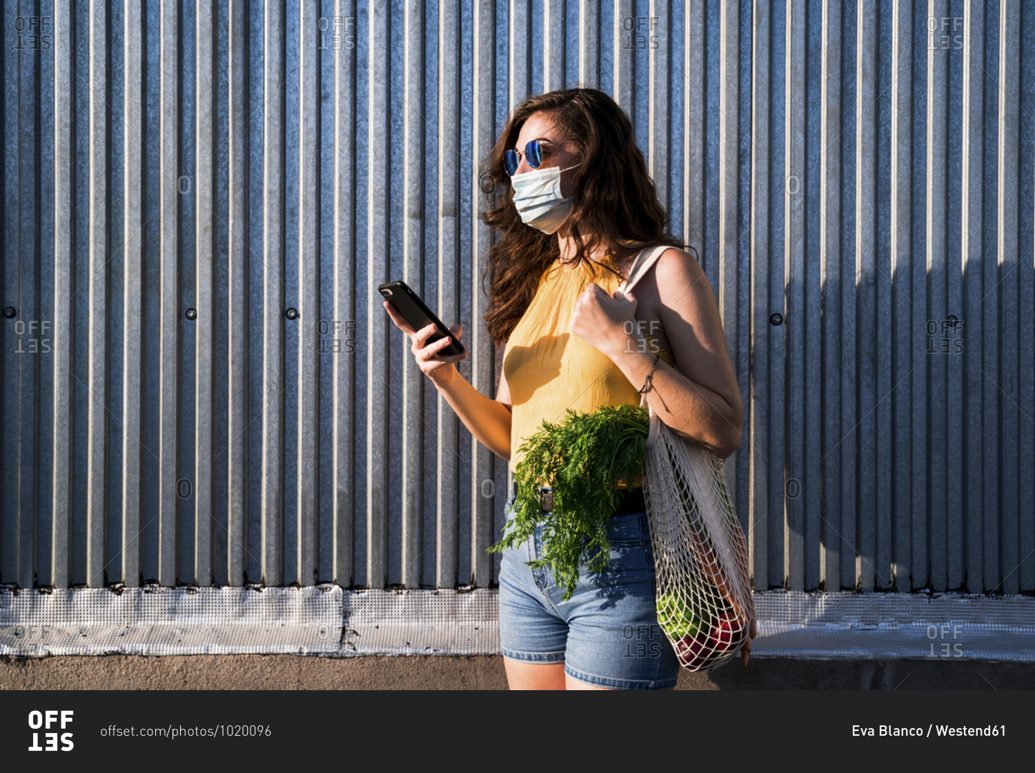 Young woman with vegetables in reusable mesh bag using smart phone while standing against metallic wall