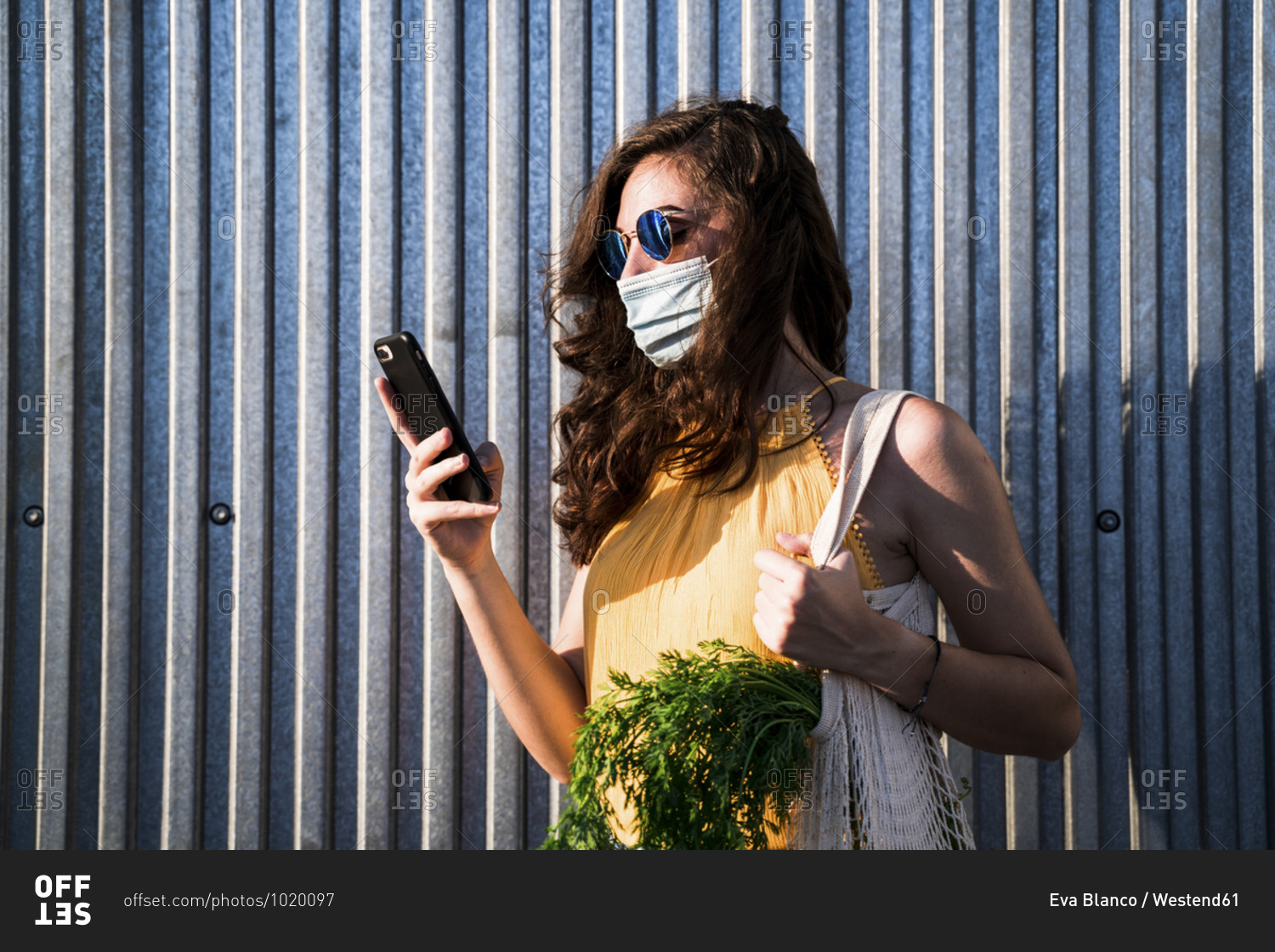 Young woman wearing face mask while standing with mobile phone and reusable mesh bag against metallic wall