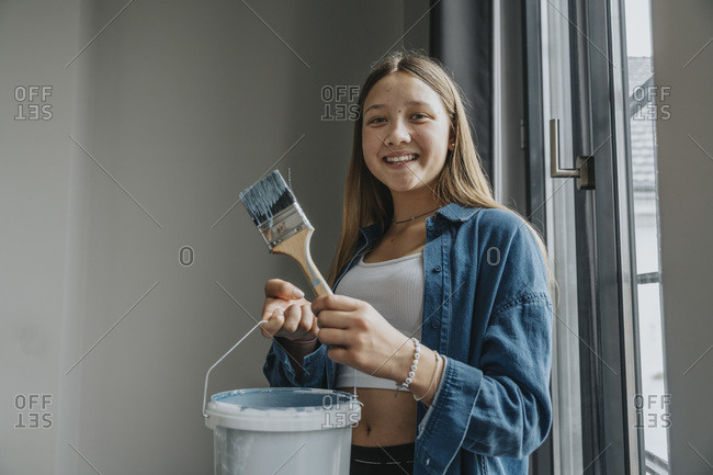 Smiling teenage girl standing in room- holding paint bucket and brush