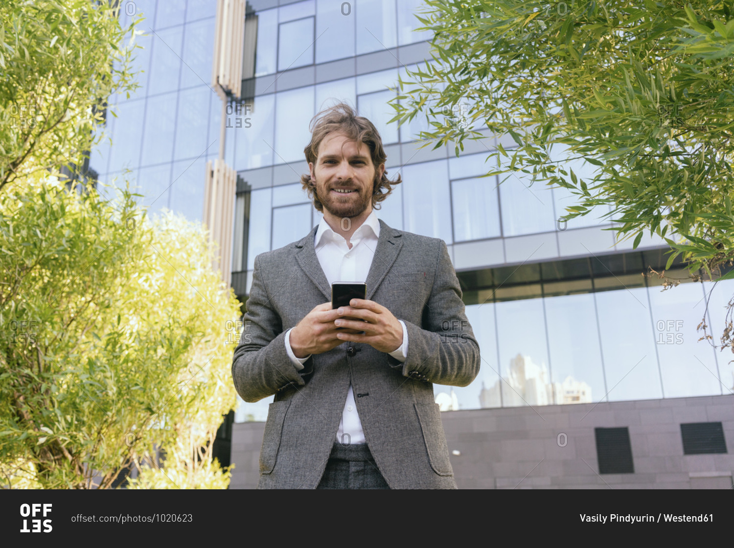 Beard young male professional with smart phone while standing against office building