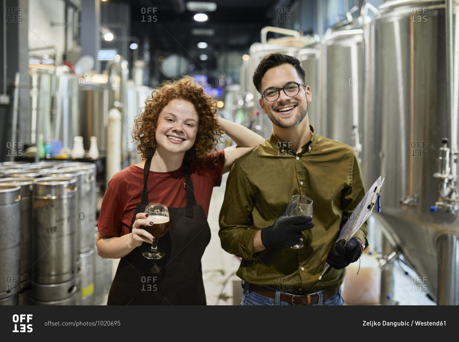 Portrait of happy man and woman holding clipboard and beer glass in craft brewery