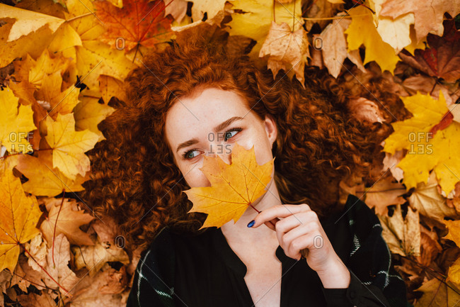 Young woman holding leaf on face lying in orange leaves in autumn park