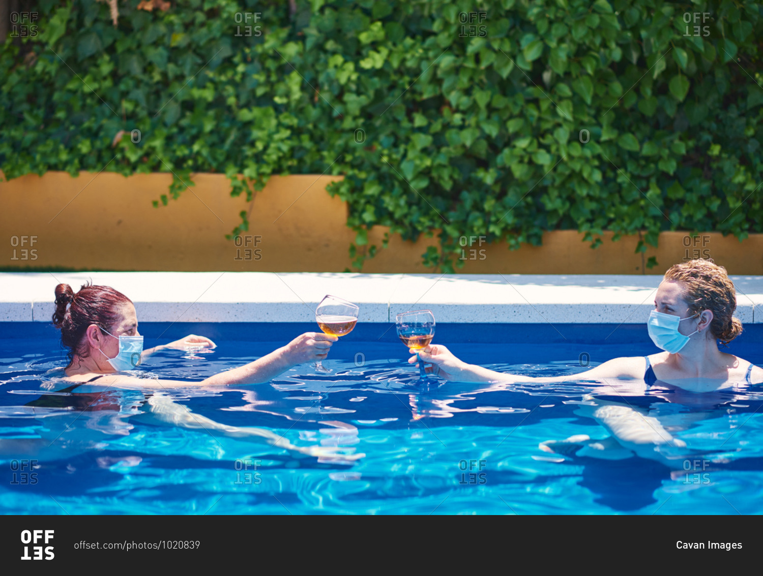 Two women toast in a pool. they wear masks to prevent the spread of the virus.