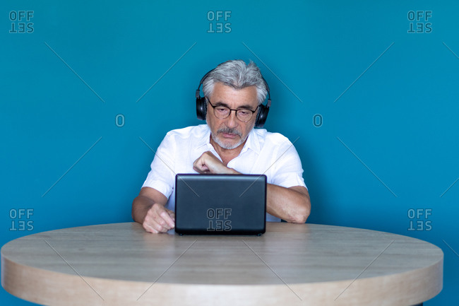 Older man with gray hair and glasses studying from home with his laptop and black headphones.