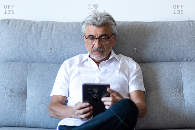 Older man with gray hair and glasses relaxing on his gray couch watching his tablet.