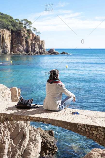 Rear view of a woman relaxing in front of blue sea, sitting on bridge
