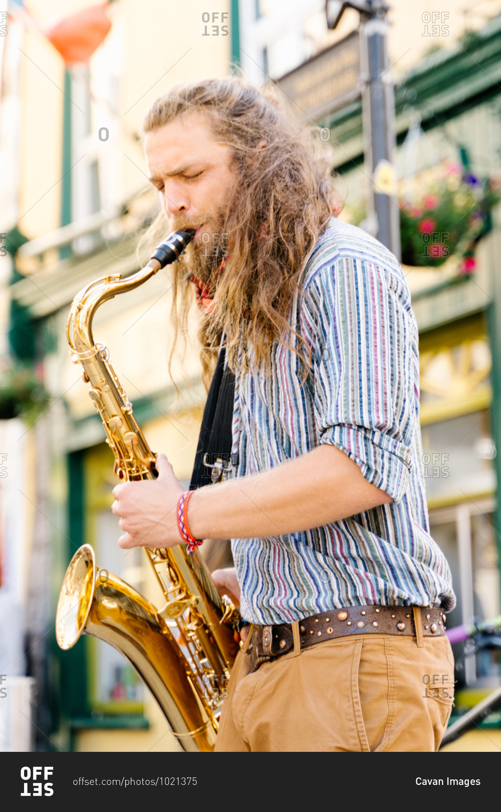Vertical photo of a young man with long hair playing saxophone in the street