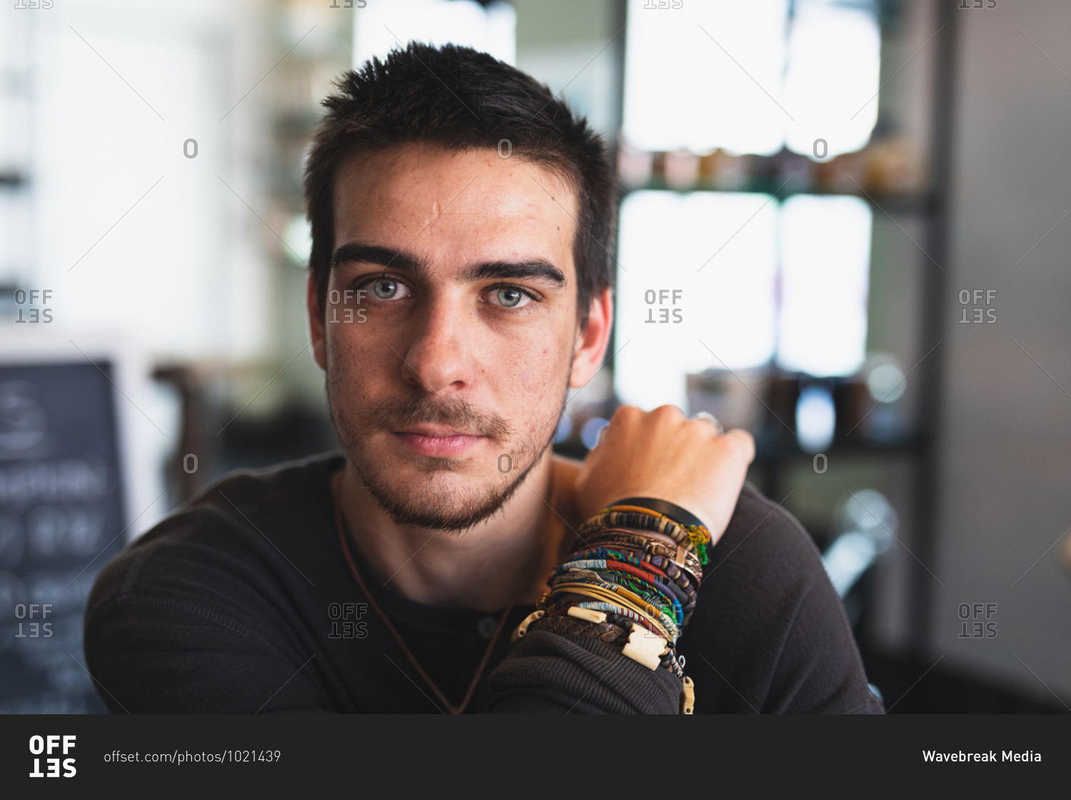 Portrait of a Caucasian man wearing casual clothes and jewelry, sitting by a table in a coffee shop, looking straight into a camera