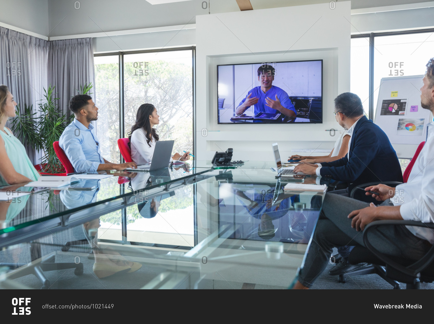 Multi-ethnic group of male and female business colleagues sitting in video conference with an Asian male colleague talking on screen. Creative business professionals working in a busy modern office.