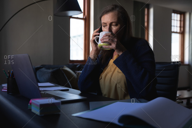 Caucasian woman enjoying time at home, social distancing and self isolation in quarantine lockdown, sitting at table, using a laptop, talking on smartphone and drinking tea.