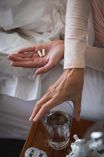 Mid section of sick Caucasian woman spending time at home, social distancing and self isolation in quarantine lockdown, lying in bed, holding pills, reaching for water.
