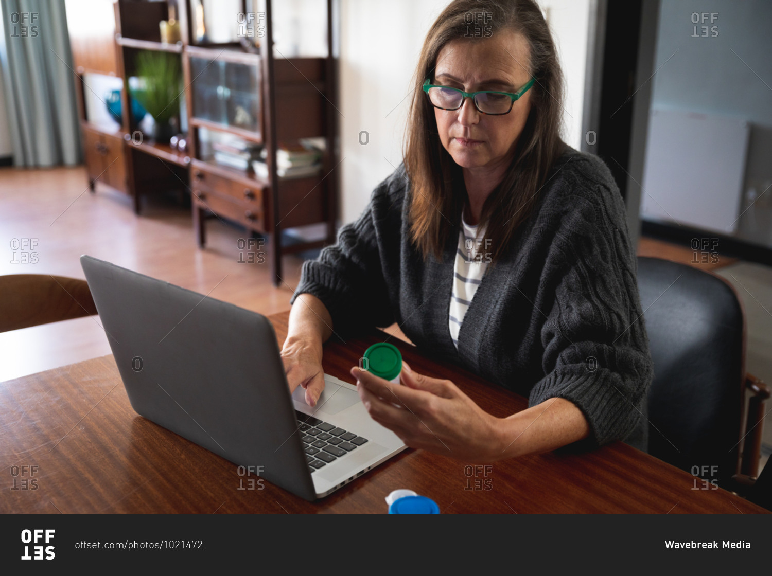 Caucasian woman enjoying time at home, social distancing and self isolation in quarantine lockdown, sitting in living room, using laptop computer, holding medication container.