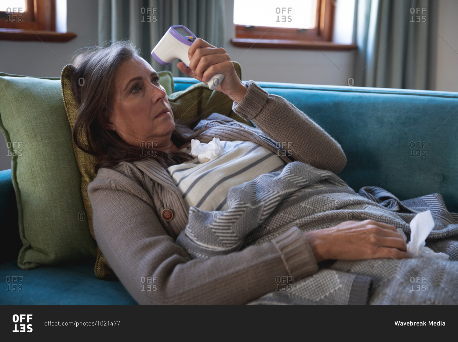 Sick Caucasian woman spending time at home, social distancing and self isolation in quarantine lockdown, lying on sofa covered with blanket, holding thermometer, measuring temperature.
