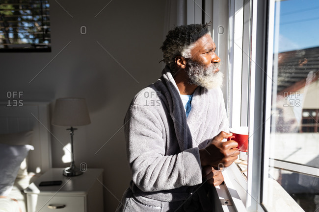 African American senior man standing in a bedroom, holding a cup of coffee, looking through a window, social distancing and self isolation in quarantine lockdown