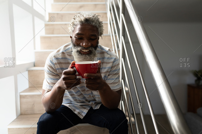 African American senior man sitting on stairs, holding a cup of coffee and smiling, social distancing and self isolation in quarantine lockdown