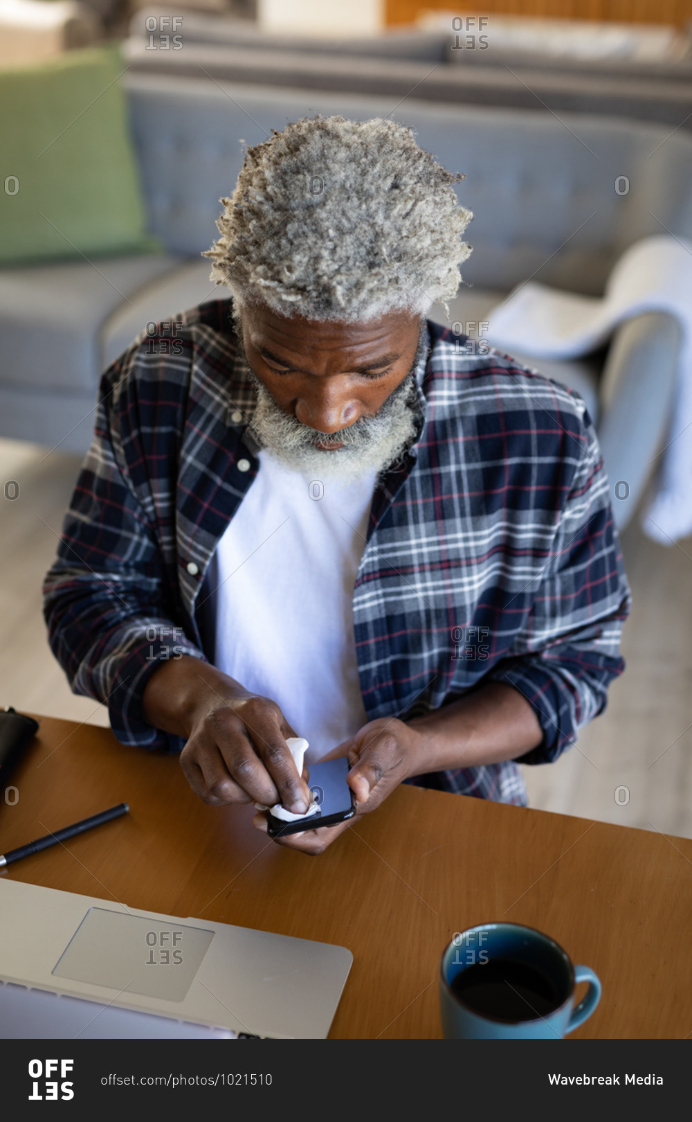 African American senior man sitting by a table, cleaning a smartphone with a tissue, social distancing and self isolation in quarantine lockdown