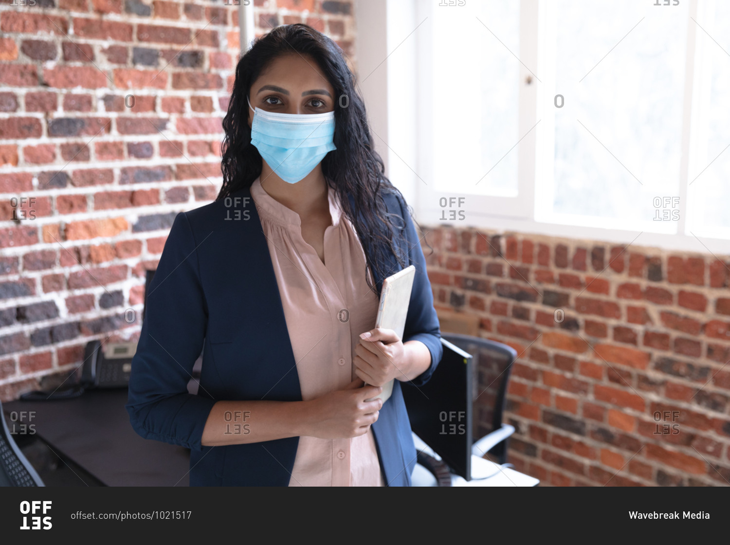 Portrait of mixed race woman working in a casual office, holding tablet, wearing face mask and looking at camera. Social distancing in the workplace during Coronavirus Covid 19 pandemic.