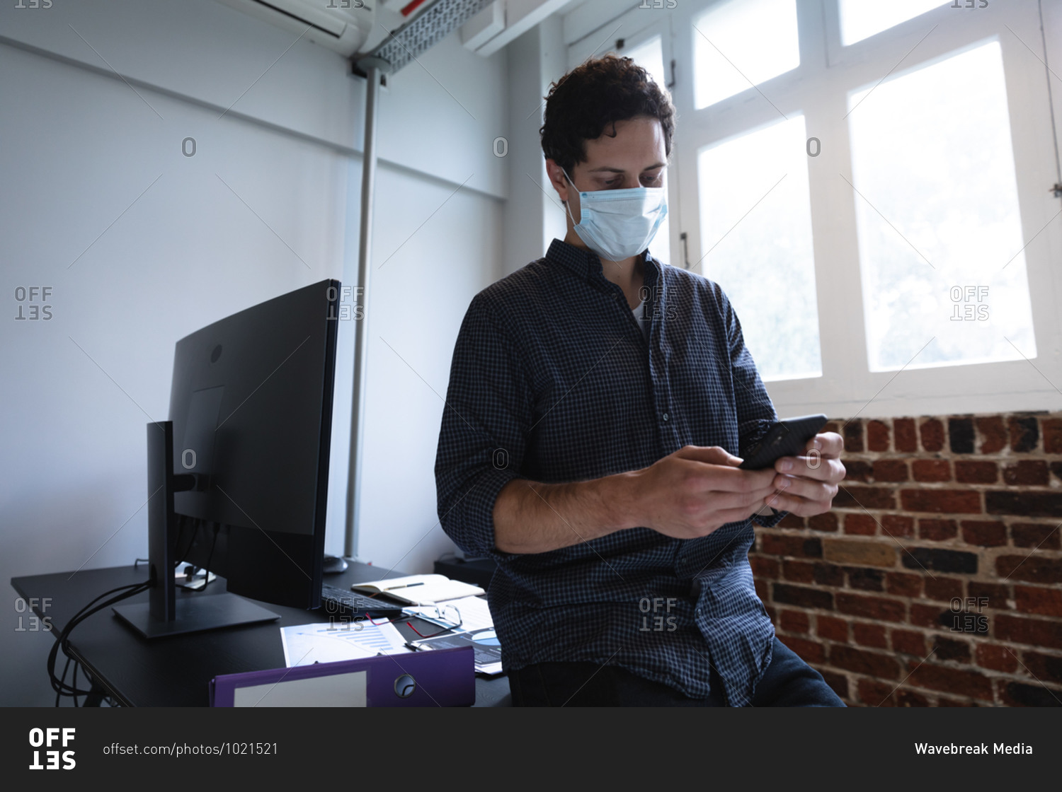Caucasian man working in a casual office, using his smartphone and wearing face mask. Social distancing in the workplace during Coronavirus Covid 19 pandemic.