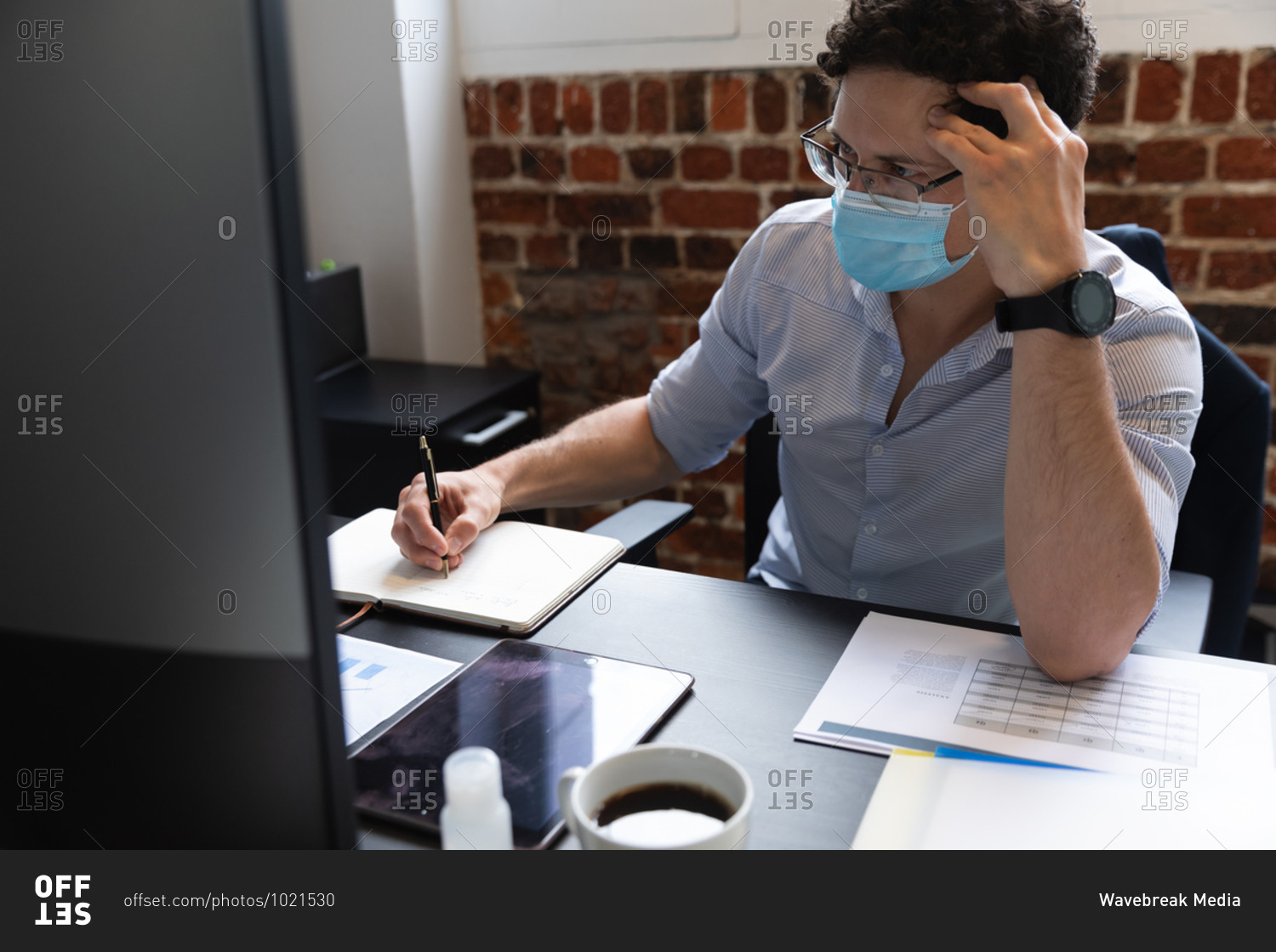 Caucasian man working in a casual office, taking notes and wearing face mask. Social distancing in the workplace during Coronavirus Covid 19 pandemic.