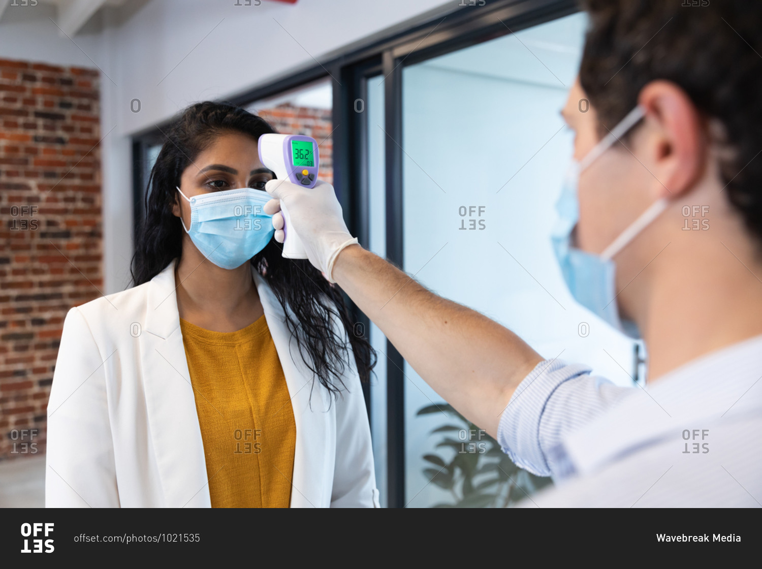 Mixed race woman and Caucasian man working in a casual office, wearing face masks, a man taking her temperature. Social distancing in the workplace during Coronavirus Covid 19 pandemic.