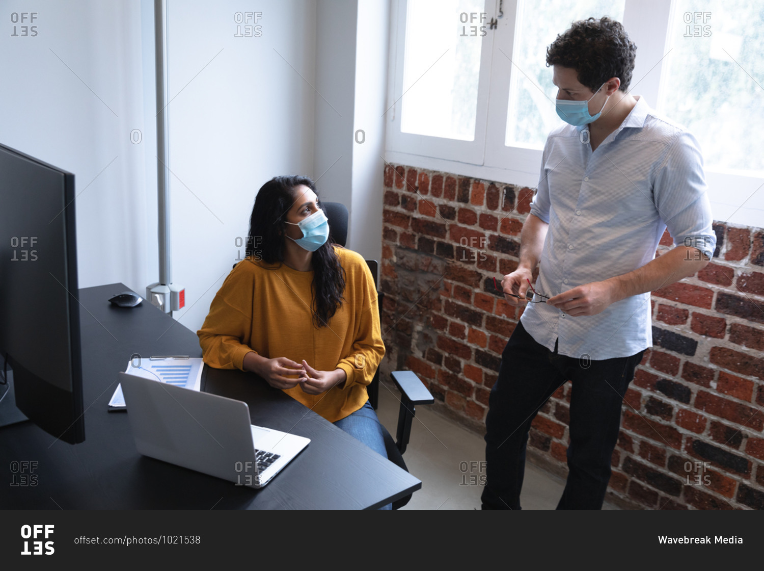 Mixed race woman and Caucasian man working in a casual office, wearing face masks and talking. Social distancing in the workplace during Coronavirus Covid 19 pandemic.