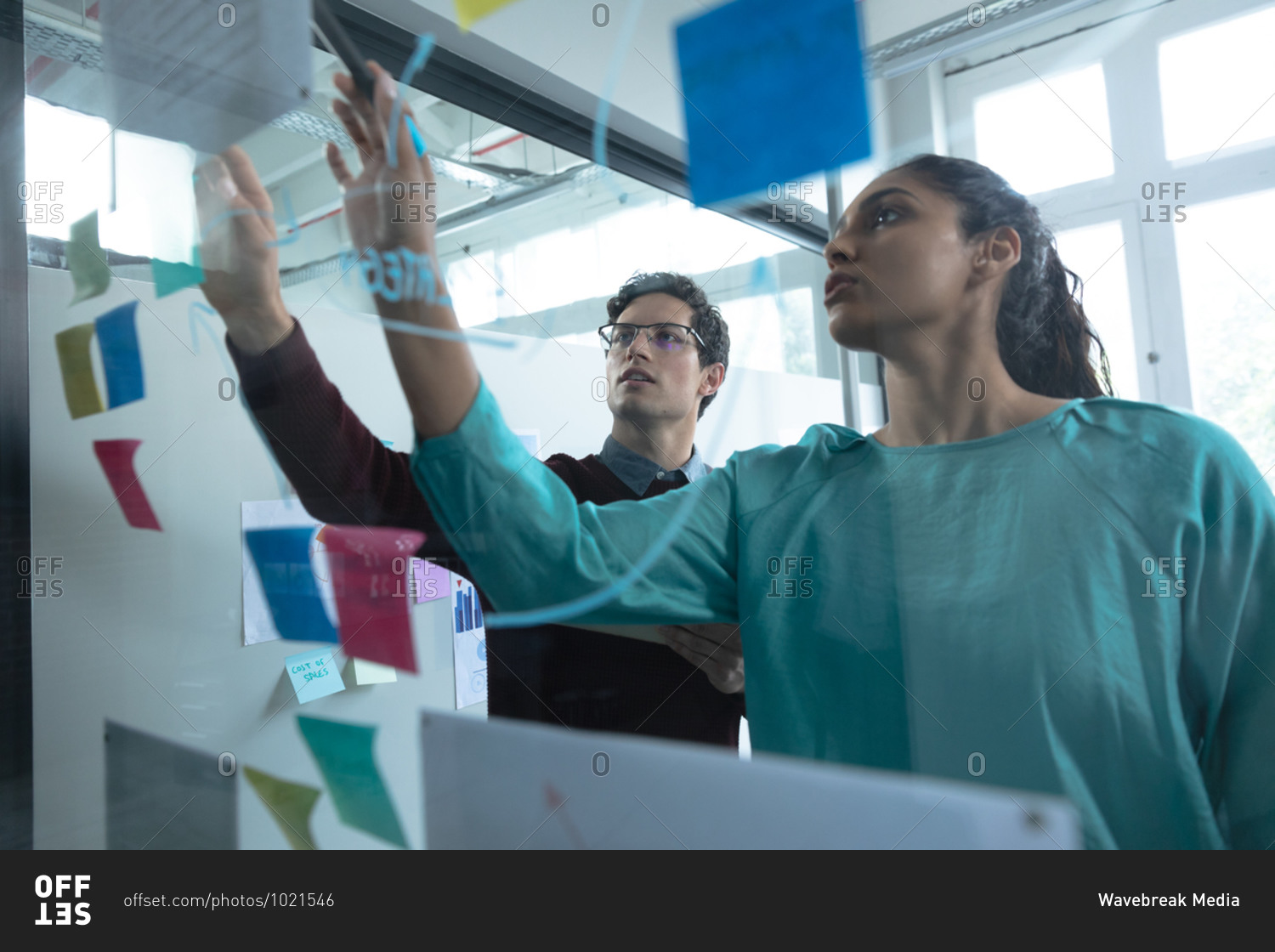 Mixed race woman and Caucasian man working in a casual office, writing on a glass board and talking. Creative business professionals working in a busy modern office.