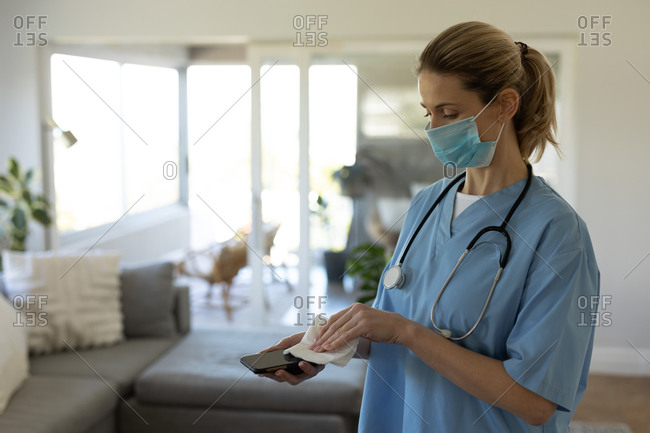 Caucasian female nurse, cleaning a smartphone with a tissue and wearing face mask. Medical care at home during Covid 19 Coronavirus quarantine.