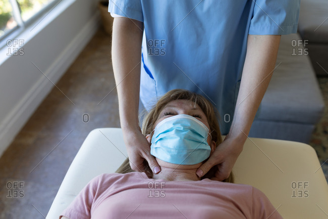 Senior Caucasian woman at home visited by Caucasian female nurse, massaging her neck, wearing face mask. Medical care
at home during Covid 19 Coronavirus quarantine.